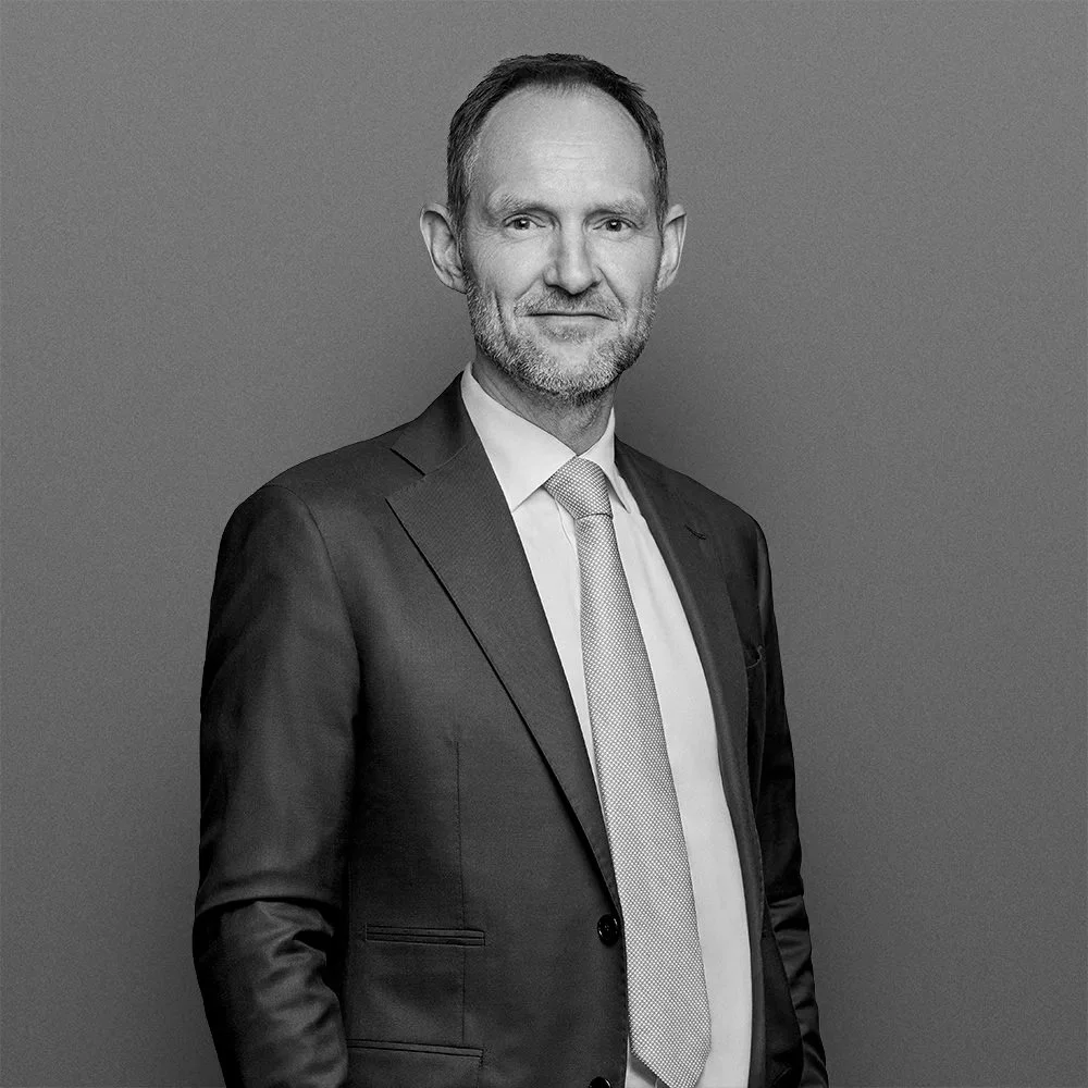 A middle-aged man with short hair and a beard, wearing a suit and tie, standing against a gray background.