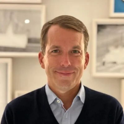 A man with short brown hair, wearing a light blue collared shirt and a navy sweater, smiling in a room with white walls and framed pictures.
