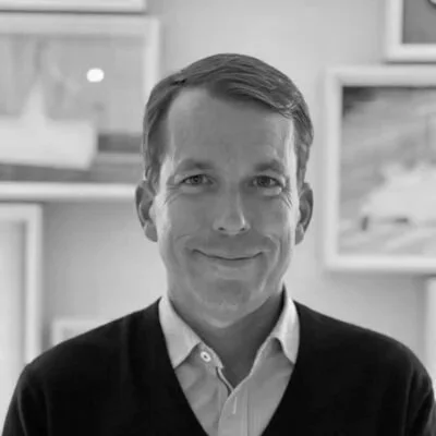 A man with short brown hair, wearing a light blue collared shirt and a navy sweater, smiling in a room with white walls and framed pictures.