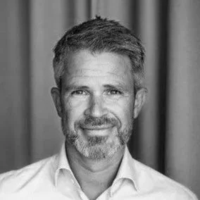 Black and white portrait of a middle-aged man with gray hair and beard, wearing a suit and white shirt, smiling, against a plain background.