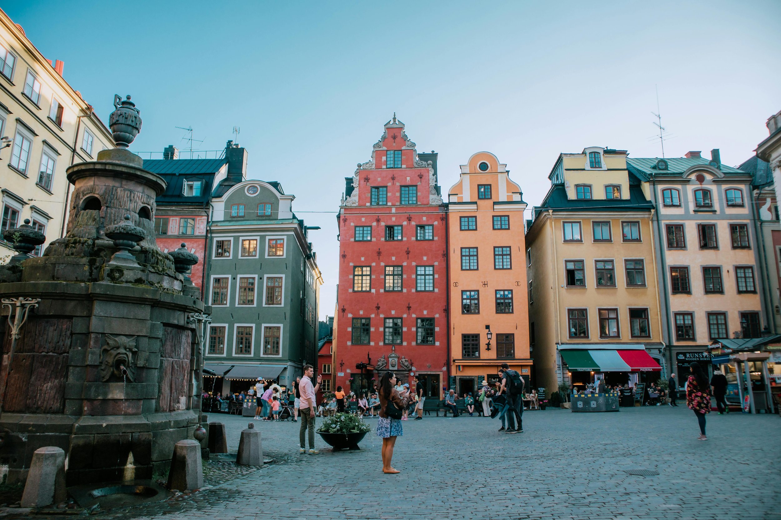 Colorful buildings lining a cobblestone square with a stone fountain in the foreground, people walking and sitting, cafes and shops on the ground floors, clear sky above.