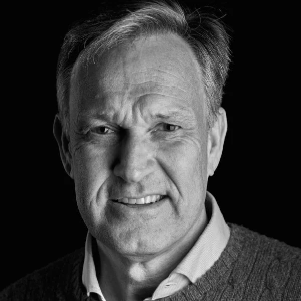 Black and white portrait of a middle-aged man with short hair, wearing a collared shirt and sweater, smiling slightly against a dark background.