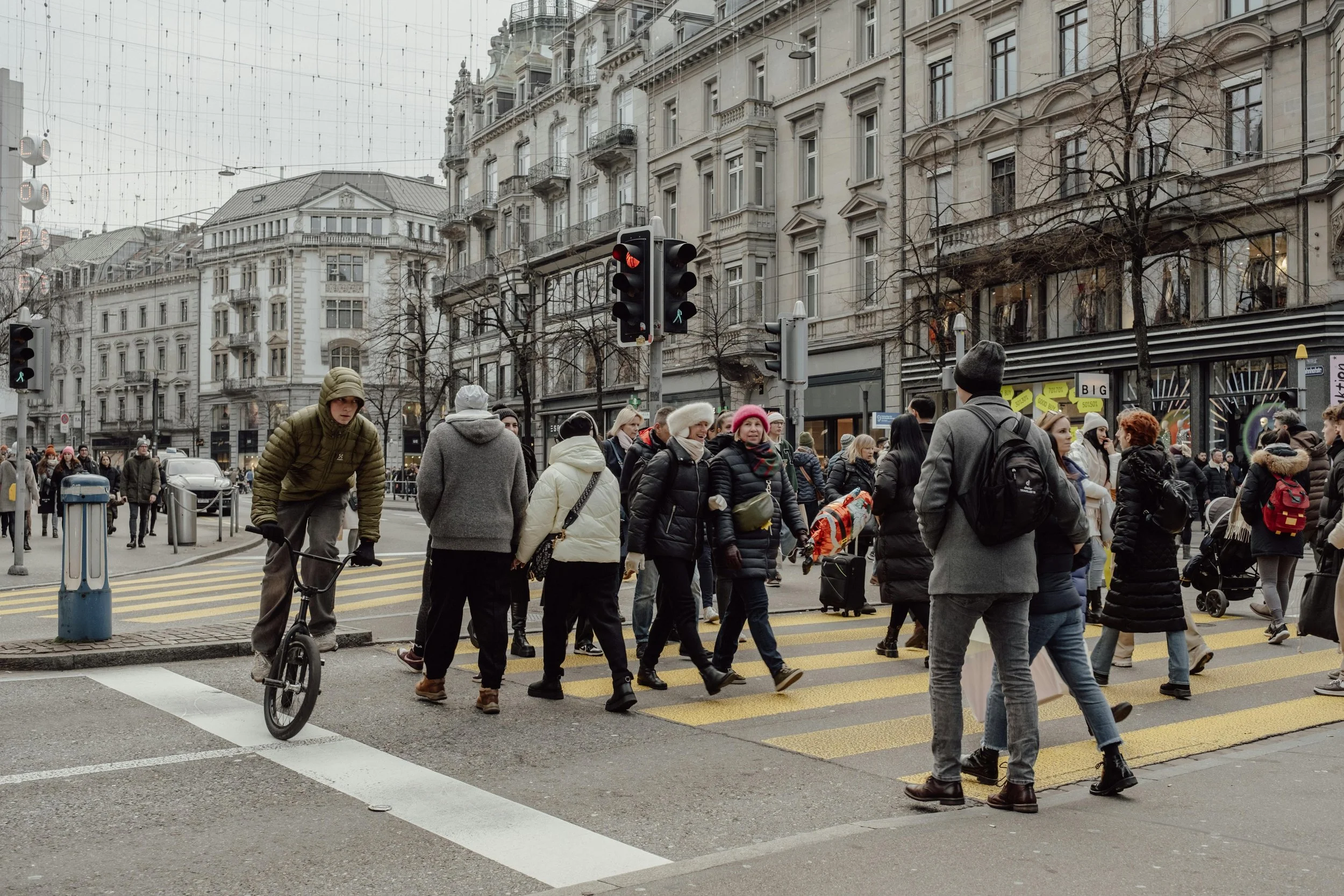 People crossing the street at a busy city intersection in winter, with classic European-style buildings in the background, and a red traffic light visible.