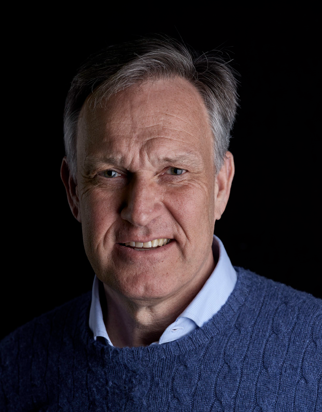 Black and white portrait of a middle-aged man with short hair, wearing a collared shirt and sweater, smiling slightly against a dark background.