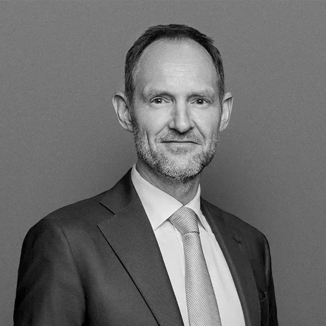 A middle-aged man with short hair and a beard, wearing a suit and tie, standing against a gray background.