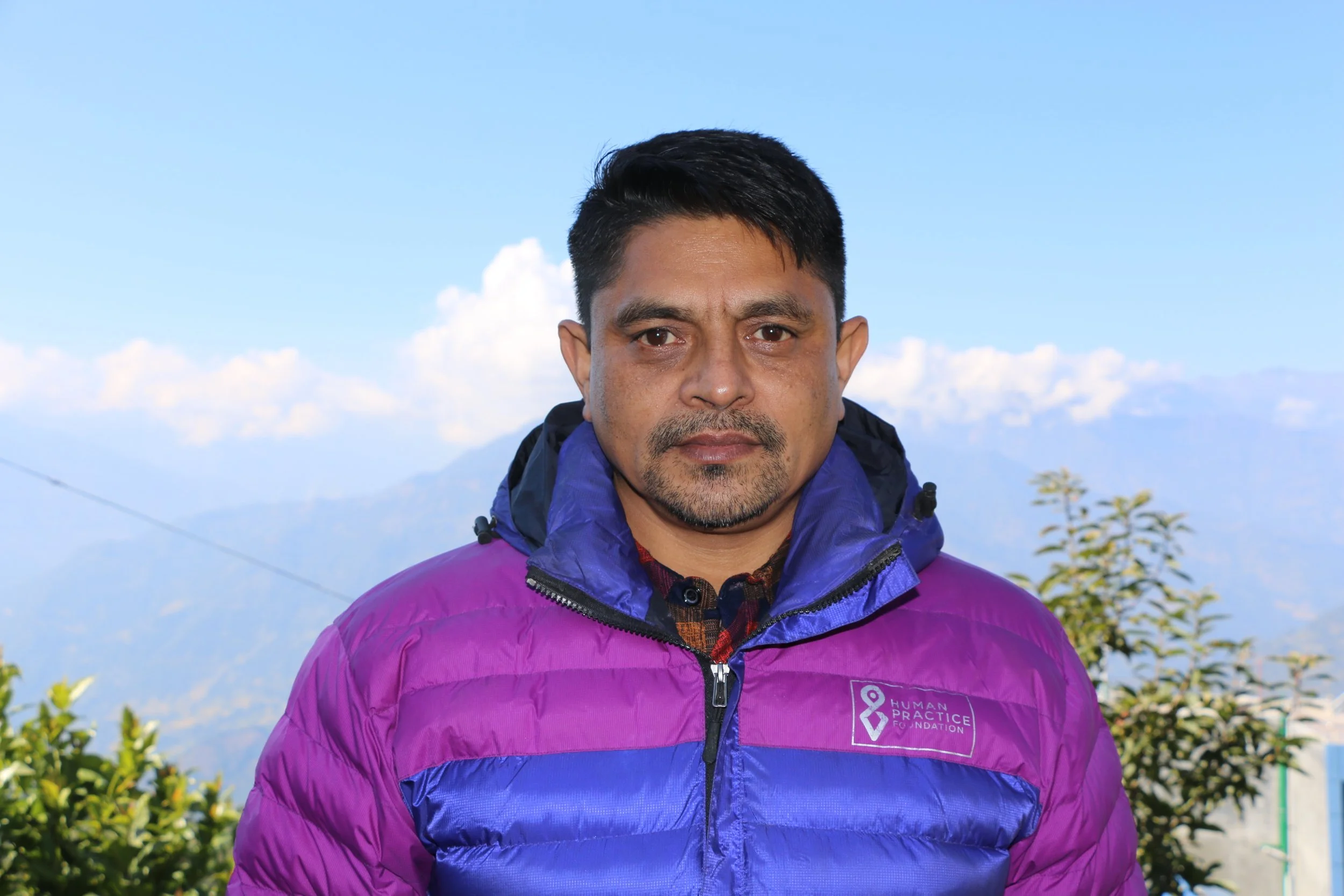 A professional man with gray hair wearing a dark suit and white shirt, standing indoors with a blurred background.