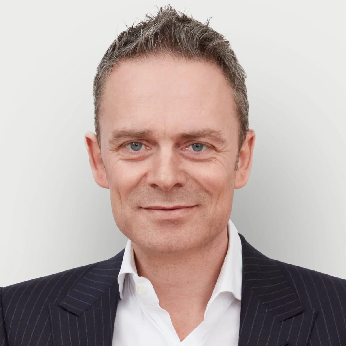 Close-up photo of a man with light skin and short, brown, possibly graying hair. He is wearing a dark, pinstripe suit and a white dress shirt, and he has a faint smile.