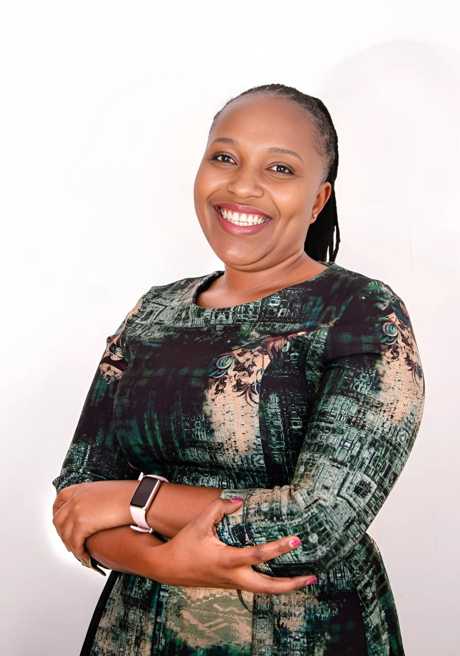 A smiling woman with braided hair, cross-armed, wearing a patterned dress and a smartwatch, standing against a white background.