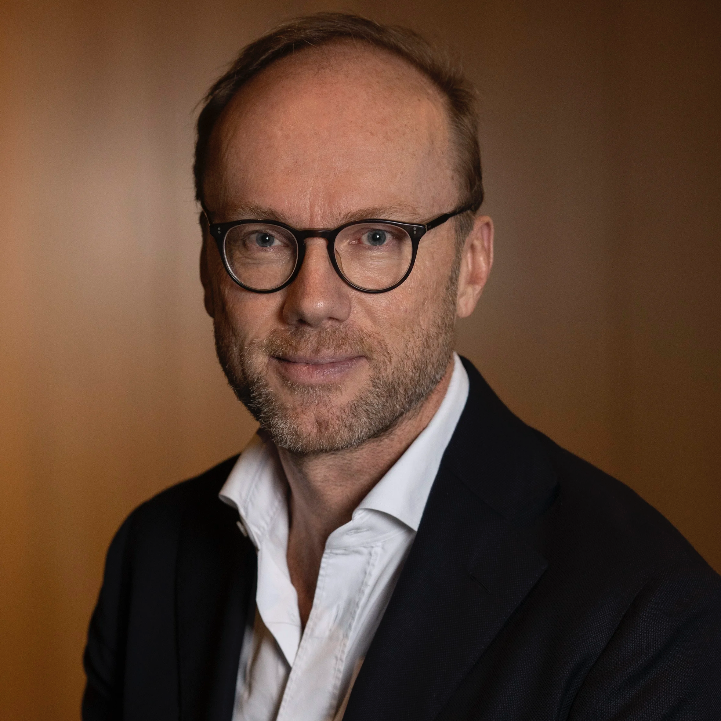 A man wearing glasses, a white shirt, and a black blazer with a blurred warm-colored background.
