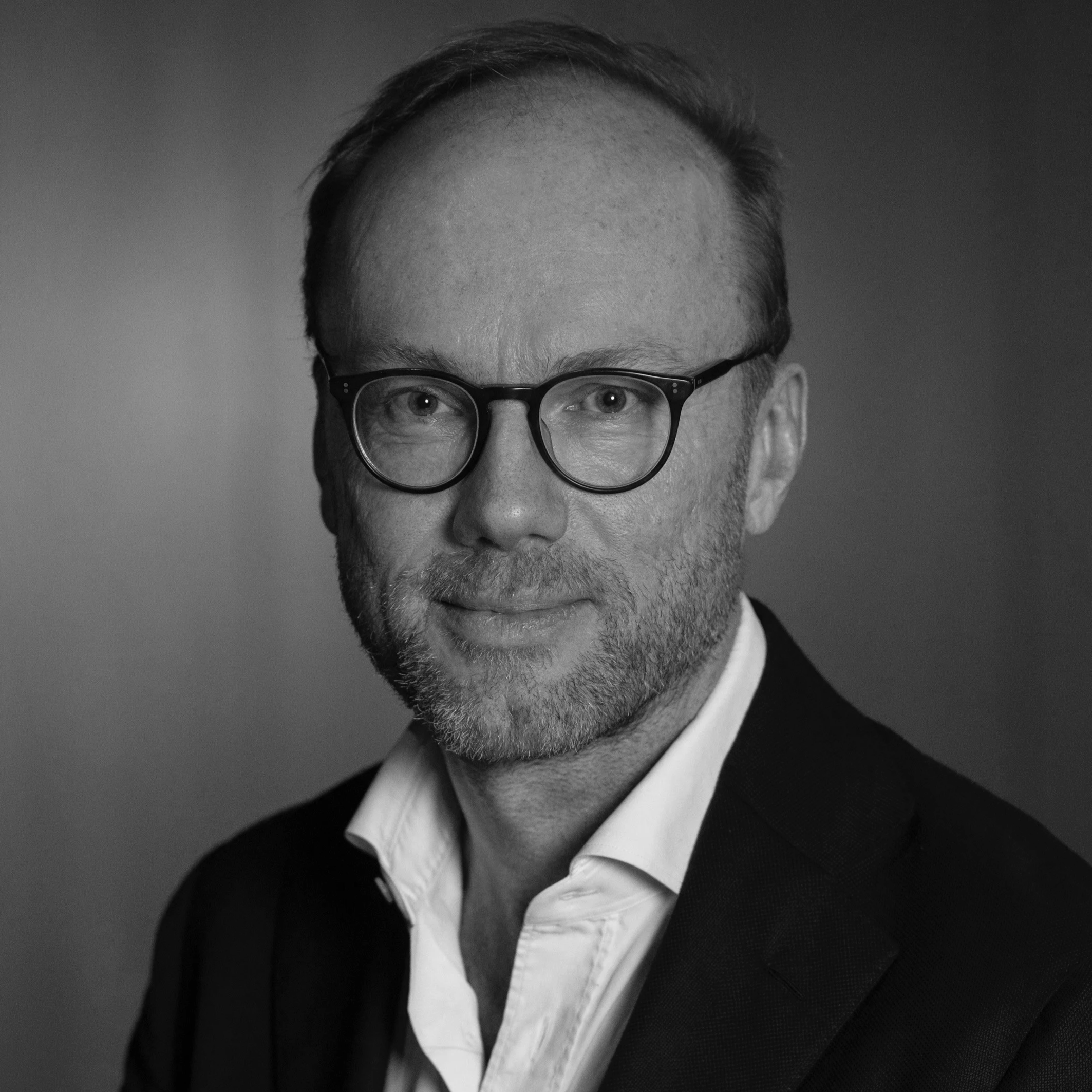 A man wearing glasses, a white shirt, and a black blazer with a blurred warm-colored background.