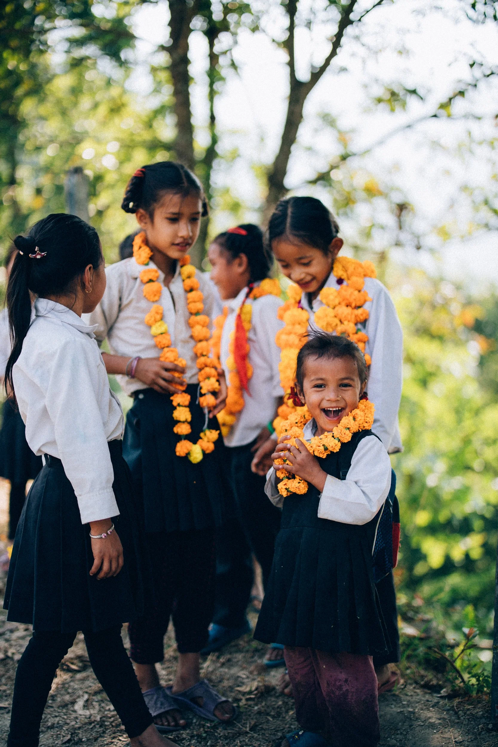 A group of young children wearing school uniforms and yellow marigold flower garlands, gathered outdoors in a forested area, smiling and engaging with each other.