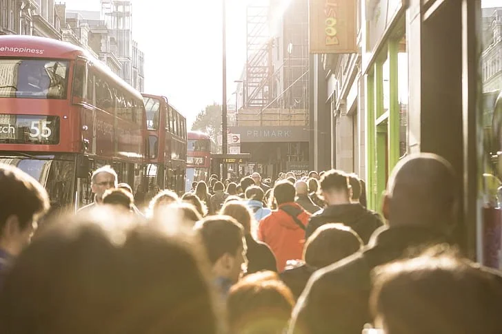 Crowded city street with people walking and buses in the background, sunlight shining through buildings.