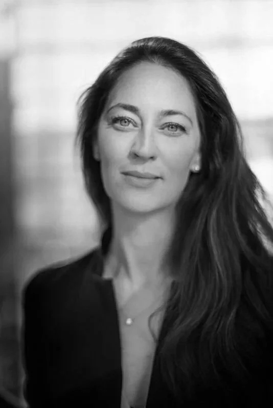 A woman with long brown hair and blue eyes posing for a portrait indoors with a blurred background.