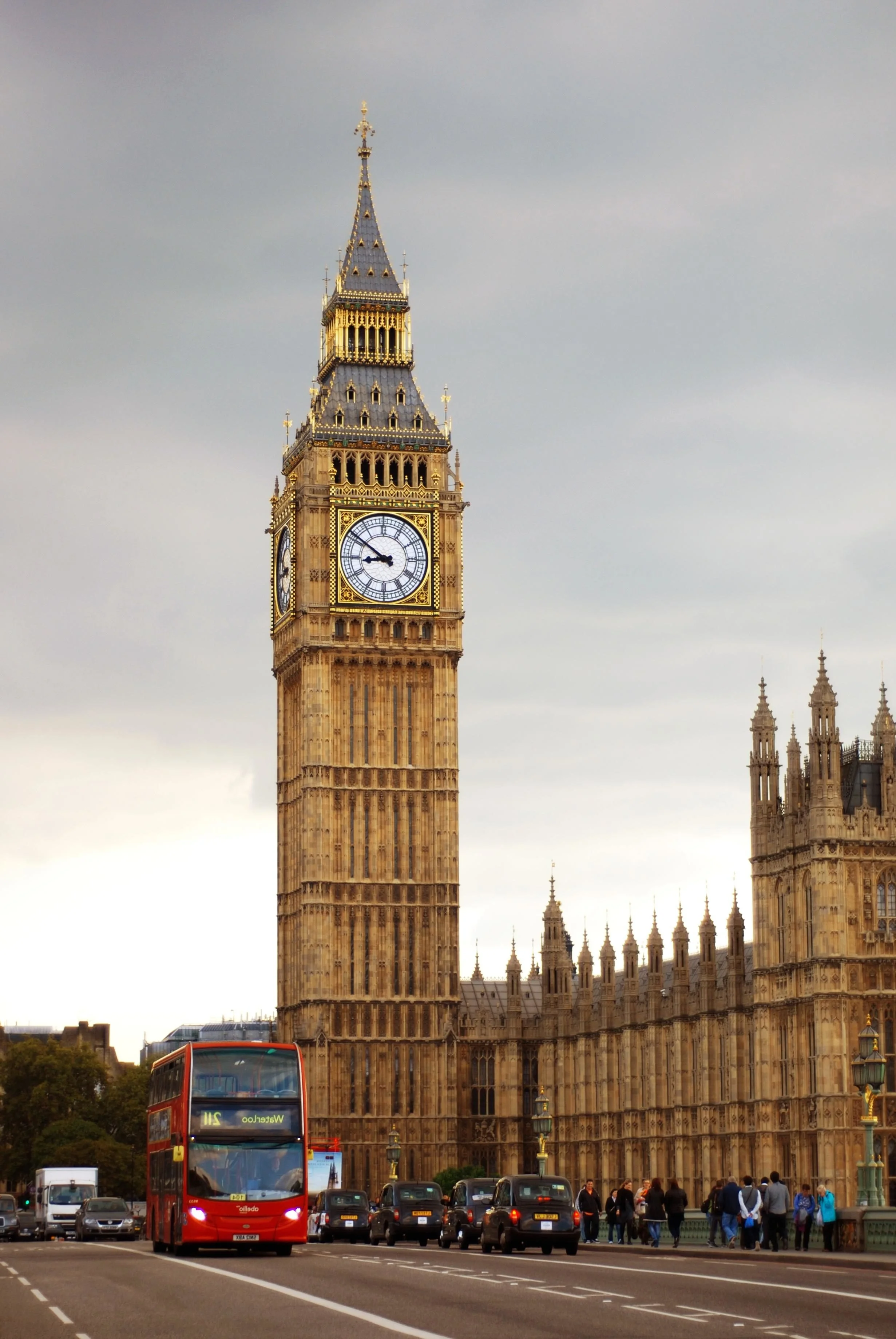 Big Ben clock tower in London with a red double-decker bus and cars on the street, and people walking near the building.