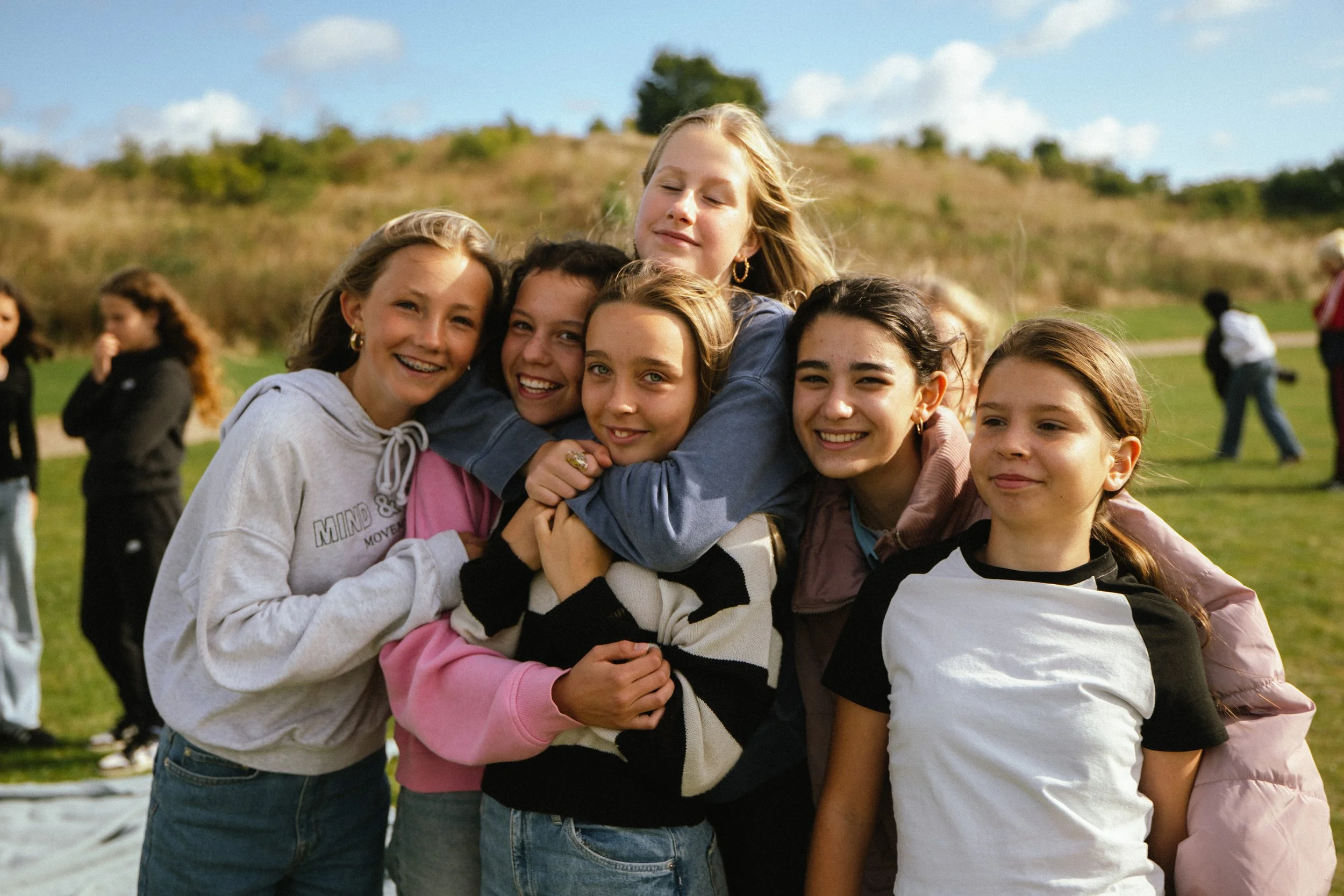 Group of six young girls hugging and smiling outdoors on a grassy field with hills and a few people in the background.