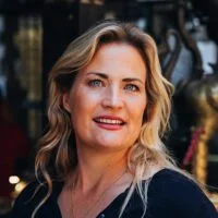 A smiling woman with wavy blonde hair, wearing a dark top, standing outdoors with trees and decorative lanterns in the background.