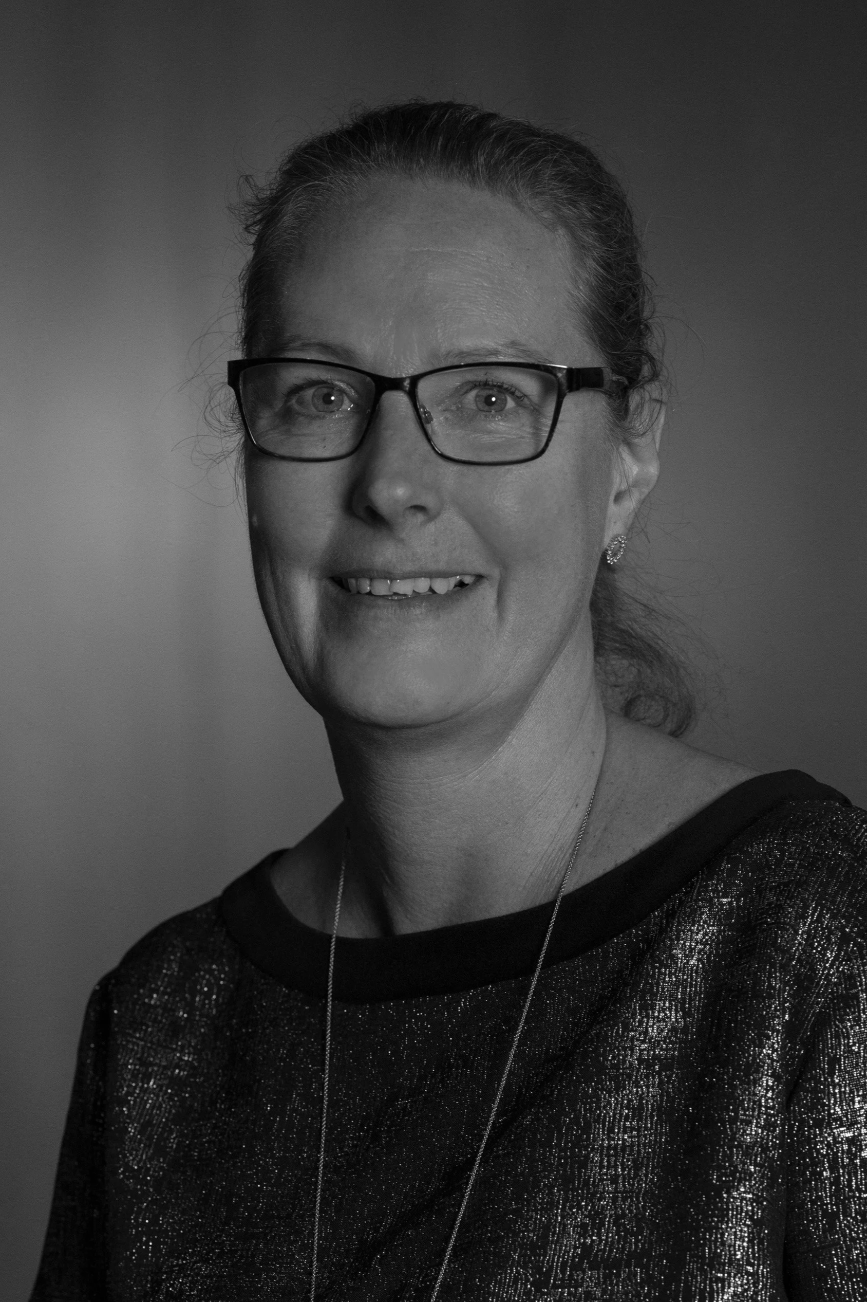 A middle-aged woman with glasses, wearing a black and gold patterned top and a necklace with a circular pendant, smiling at the camera.