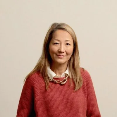 A woman with long blonde hair wearing a red sweater and a white collared shirt, standing against a plain off-white wall.