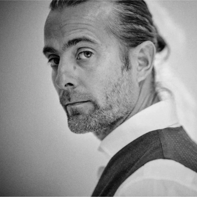 A close-up of a man with gray hair, blue eyes, and a beard, looking over his shoulder at the camera, wearing a white shirt and dark vest.