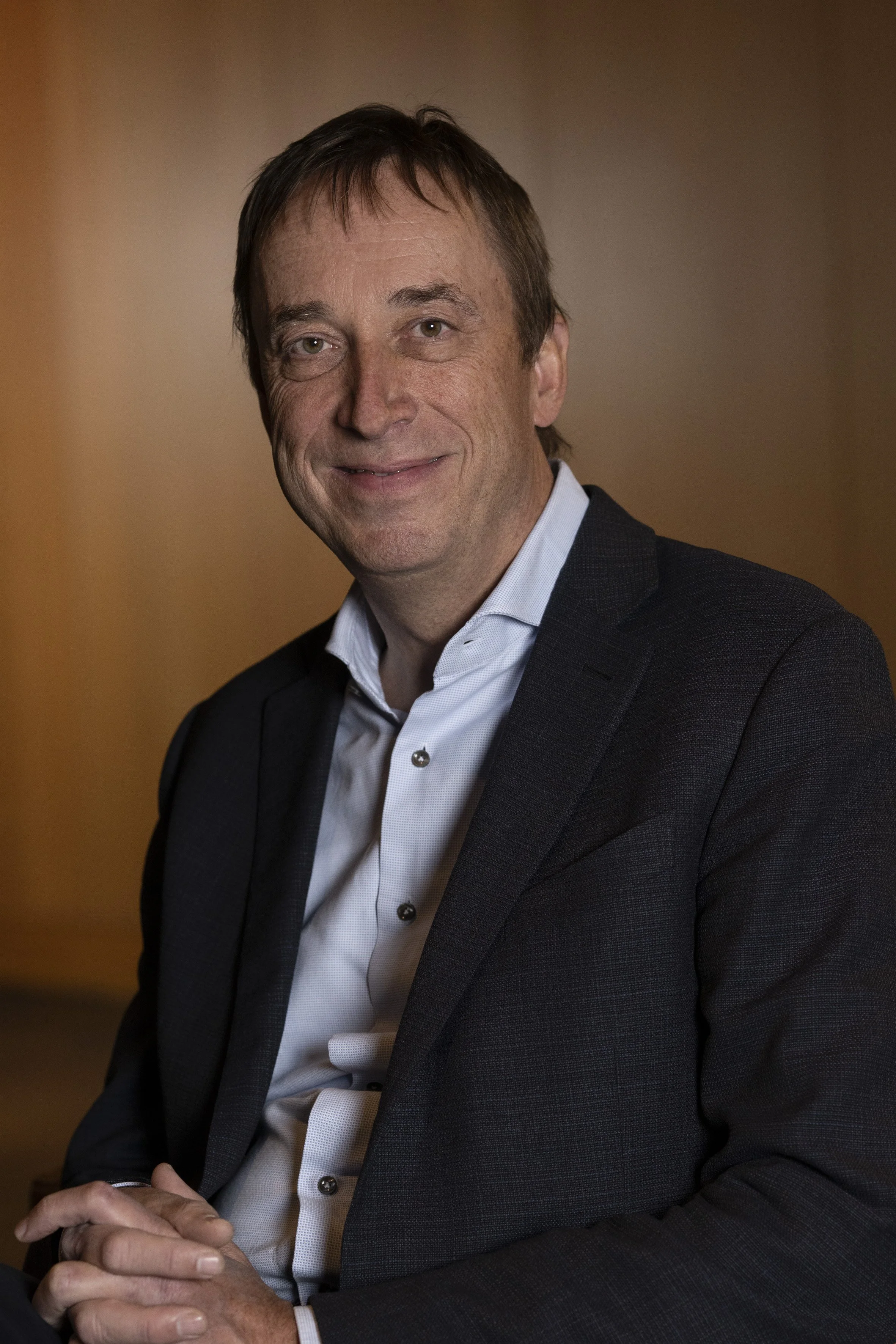 A middle-aged man with short brown hair, wearing a dark blazer and white shirt, sitting against a brown wall.