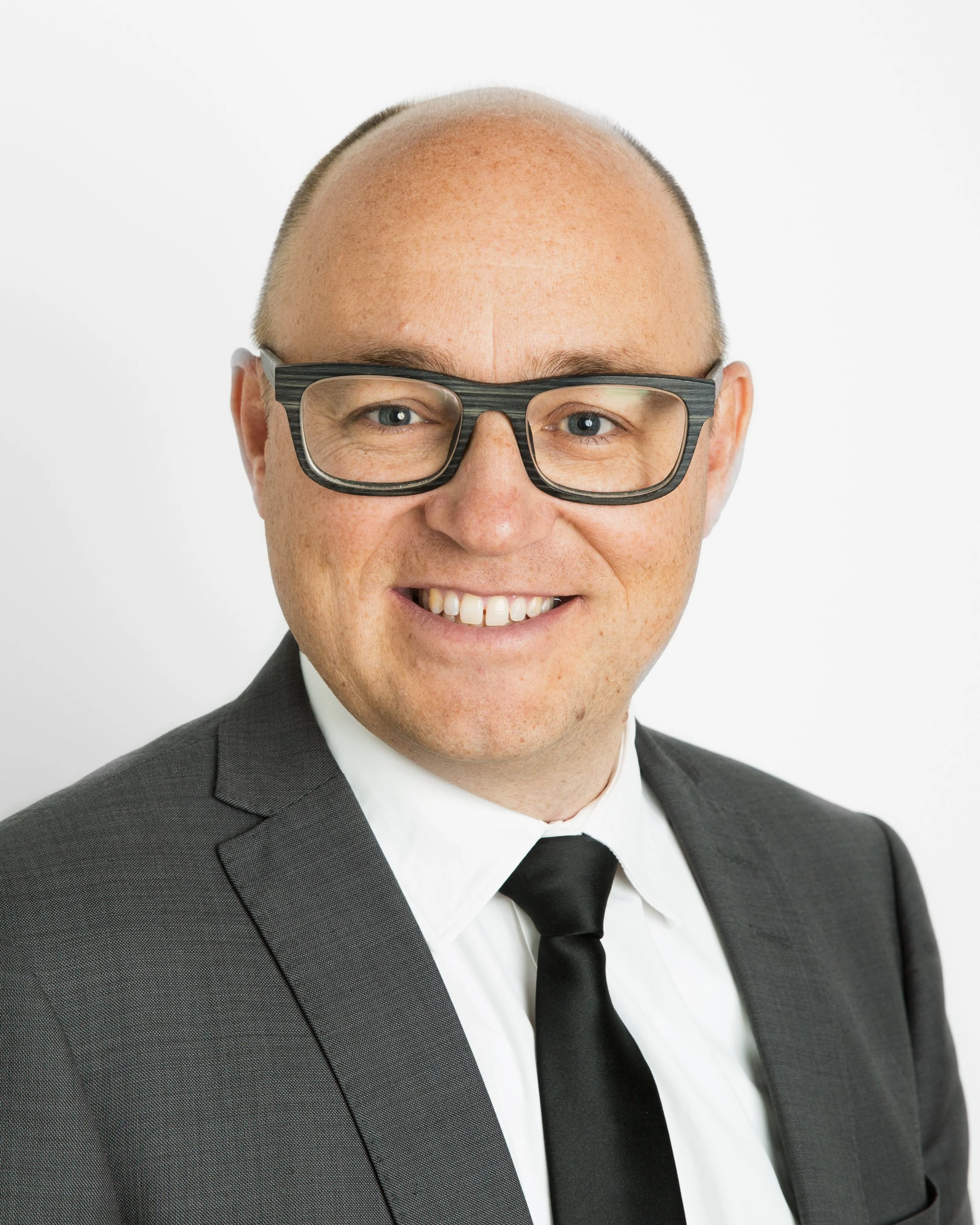 A man with glasses wearing a black suit, white shirt, and black tie, smiling against a plain white background.