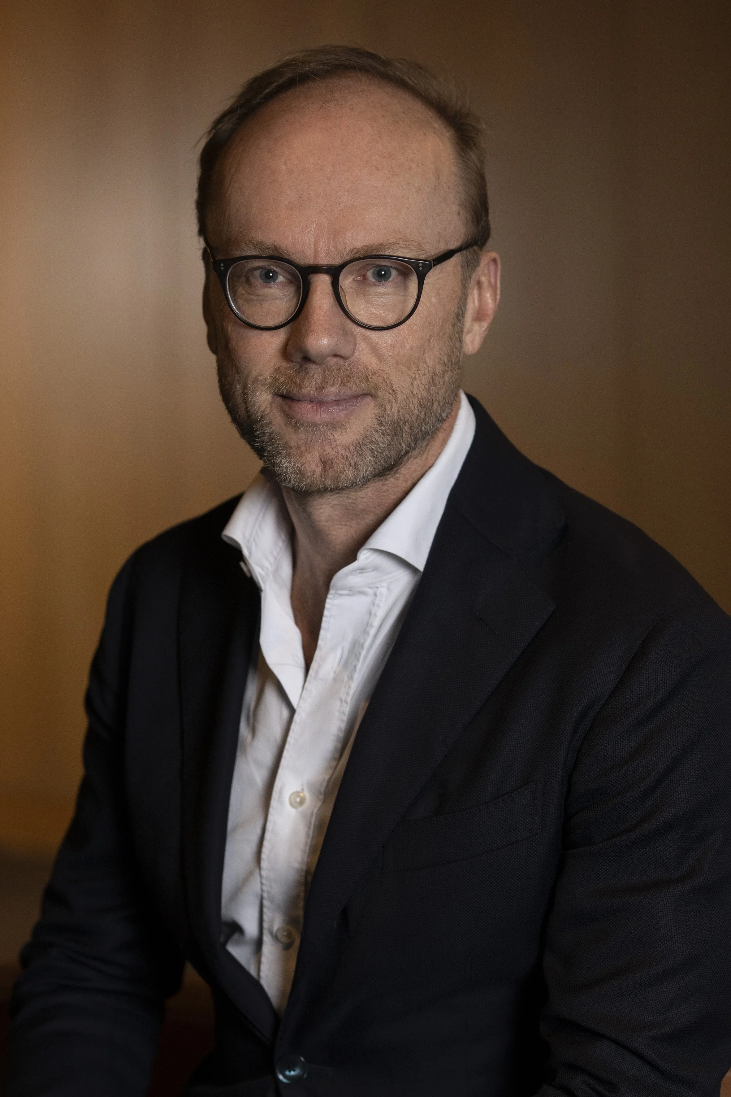 A middle-aged man with short hair and a beard, wearing a suit and tie, standing against a gray background.