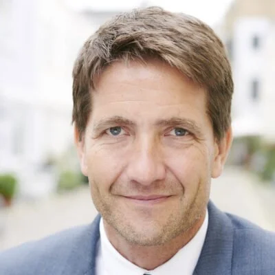 A professional man with gray hair wearing a dark suit and white shirt, standing indoors with a blurred background.