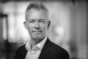 A professional man with gray hair wearing a dark suit and white shirt, standing indoors with a blurred background.