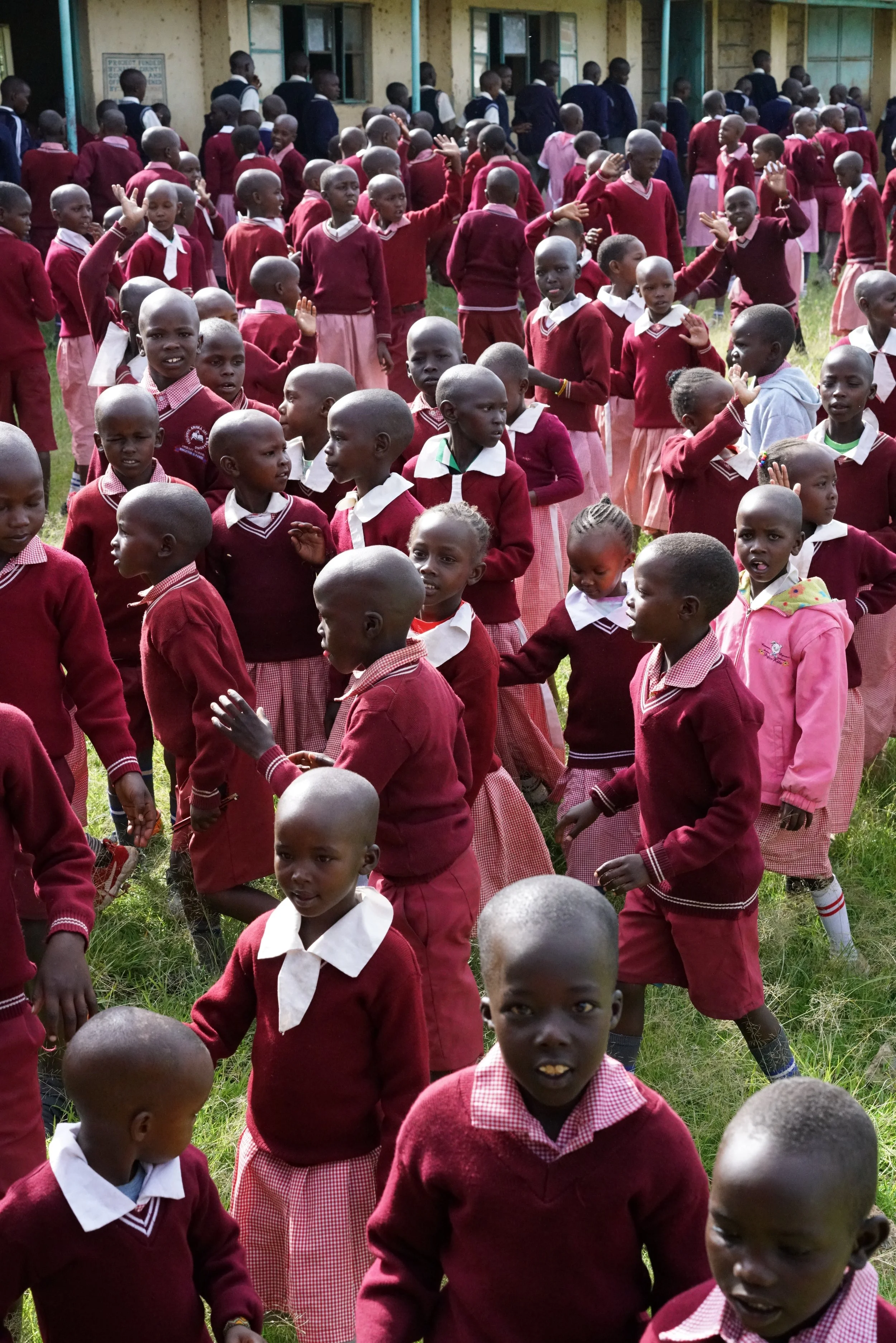 A large group of children in school uniforms are gathered outside, some are waving and talking, with a school building in the background.