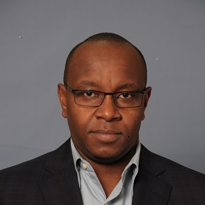 A man in a dark suit jacket and glasses staring at the camera, with a neutral expression, in front of a plain gray background.