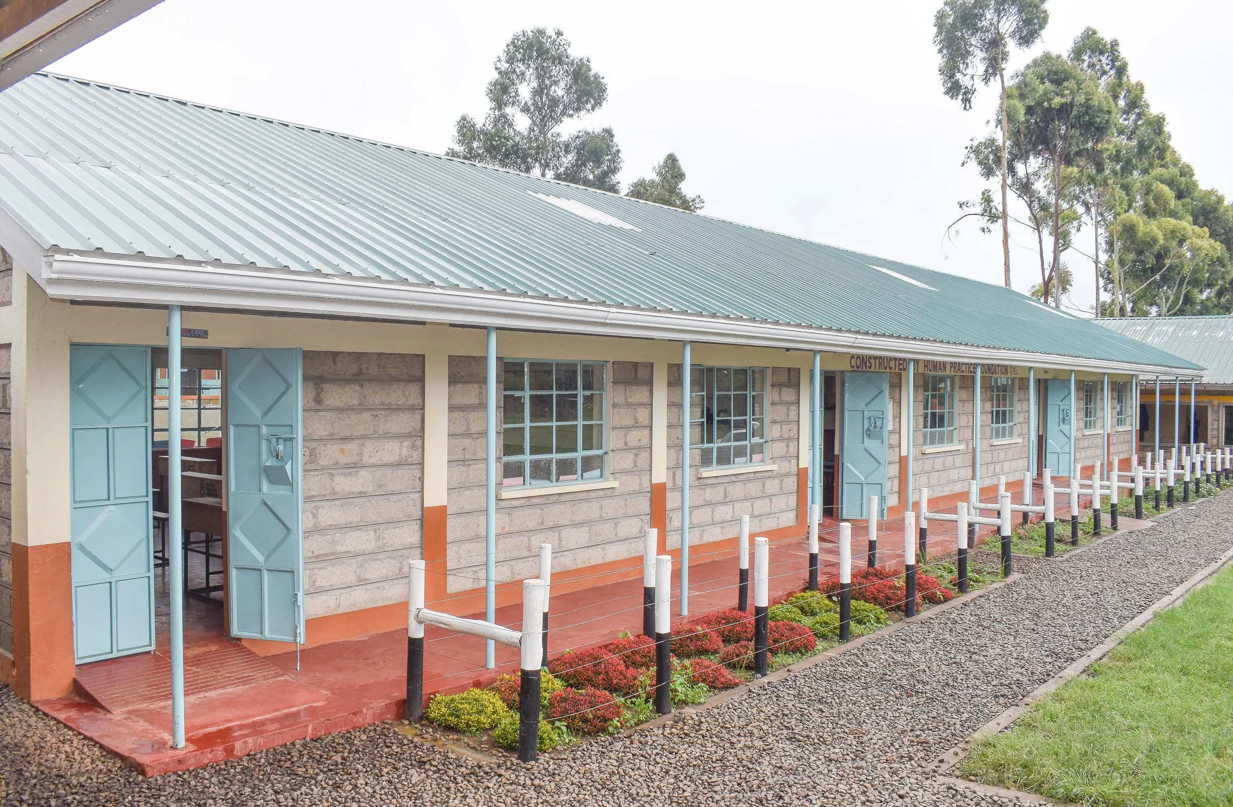 A long, single-story building with a green metal roof and light blue doors and windows. The building's exterior has brick and painted sections, with a small garden and a gravel pathway in front.