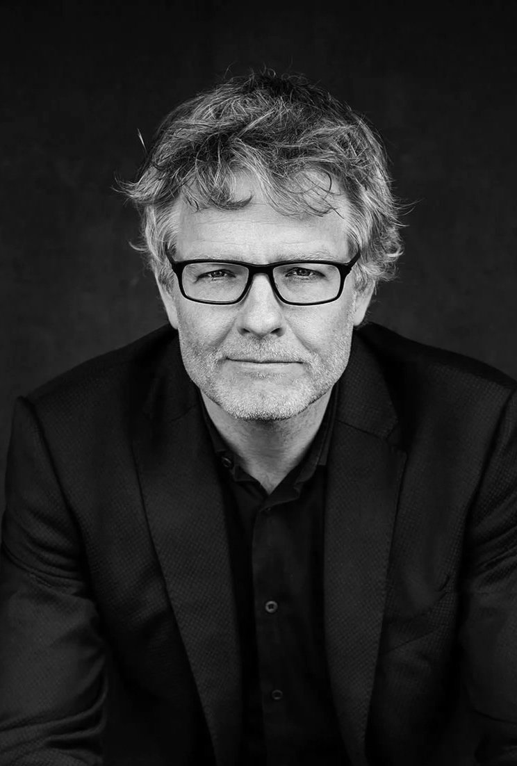A middle-aged man with gray hair and glasses, wearing a black blazer and black shirt, sitting with hands clasped, against a dark background.