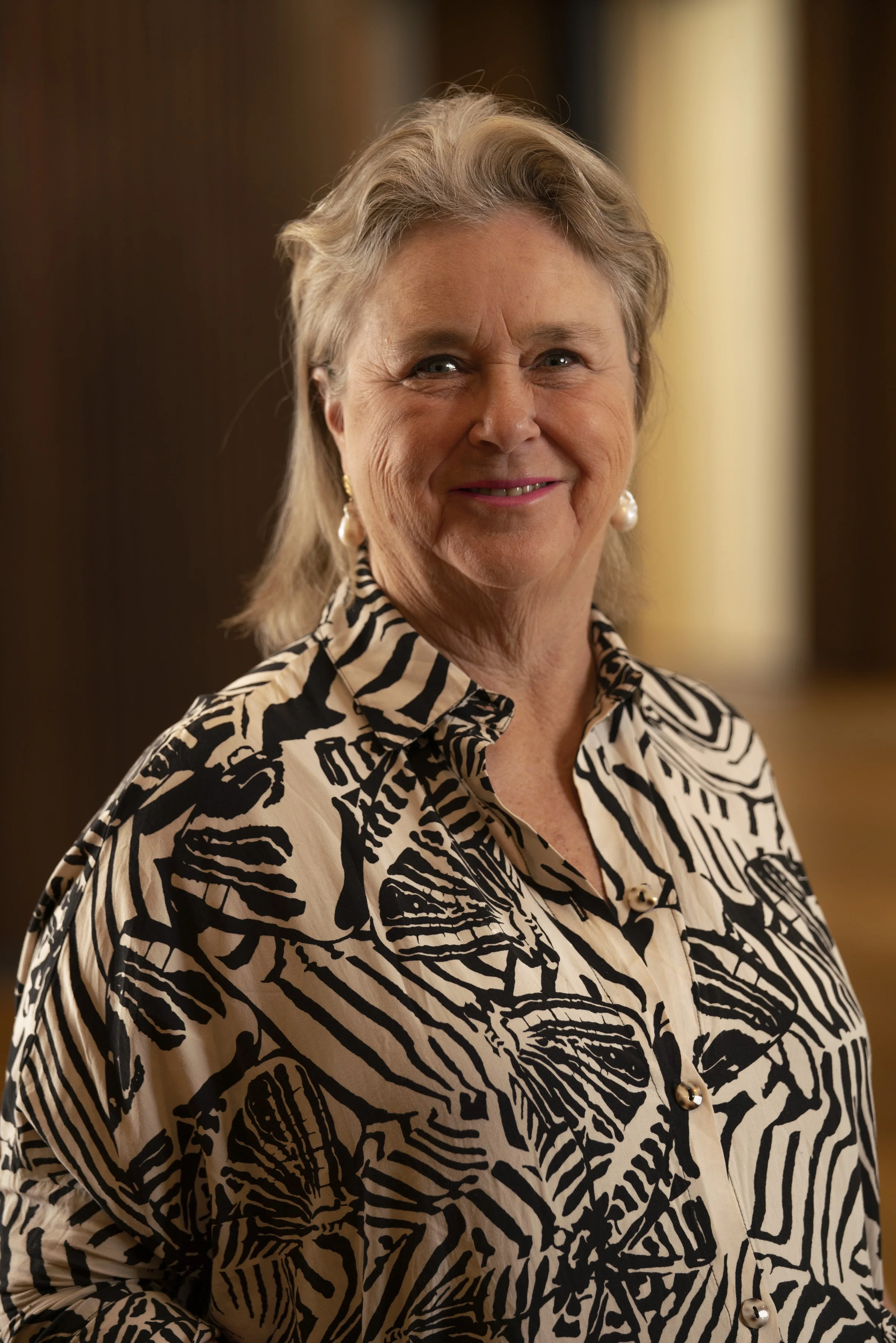 An older woman with blonde hair, wearing a black and white patterned blouse with butterfly and zebra designs, smiling with pearl earrings, standing indoors.