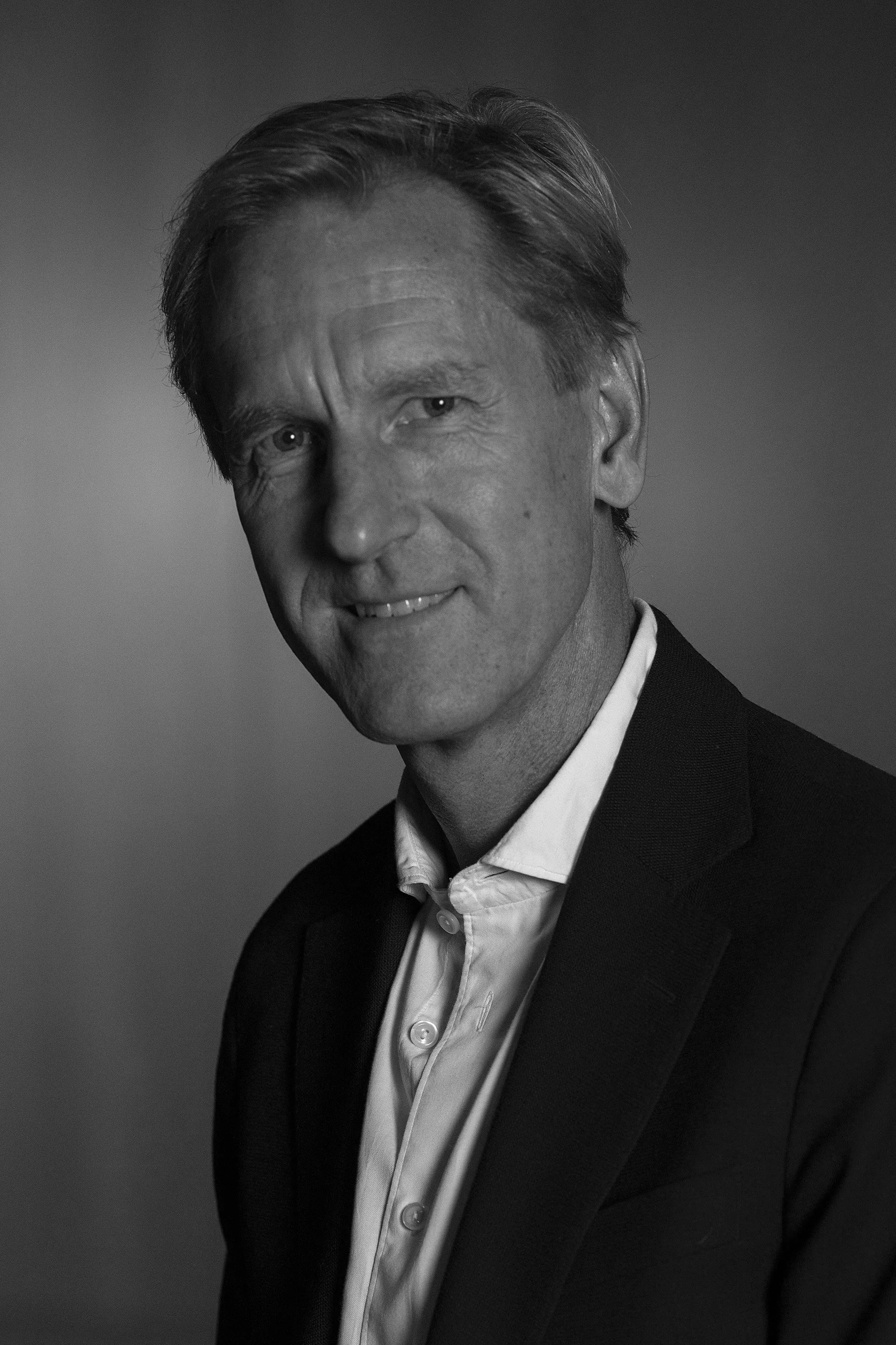A middle-aged man with blonde hair, wearing a black suit jacket and a white dress shirt, smiling slightly against a warm-colored background.