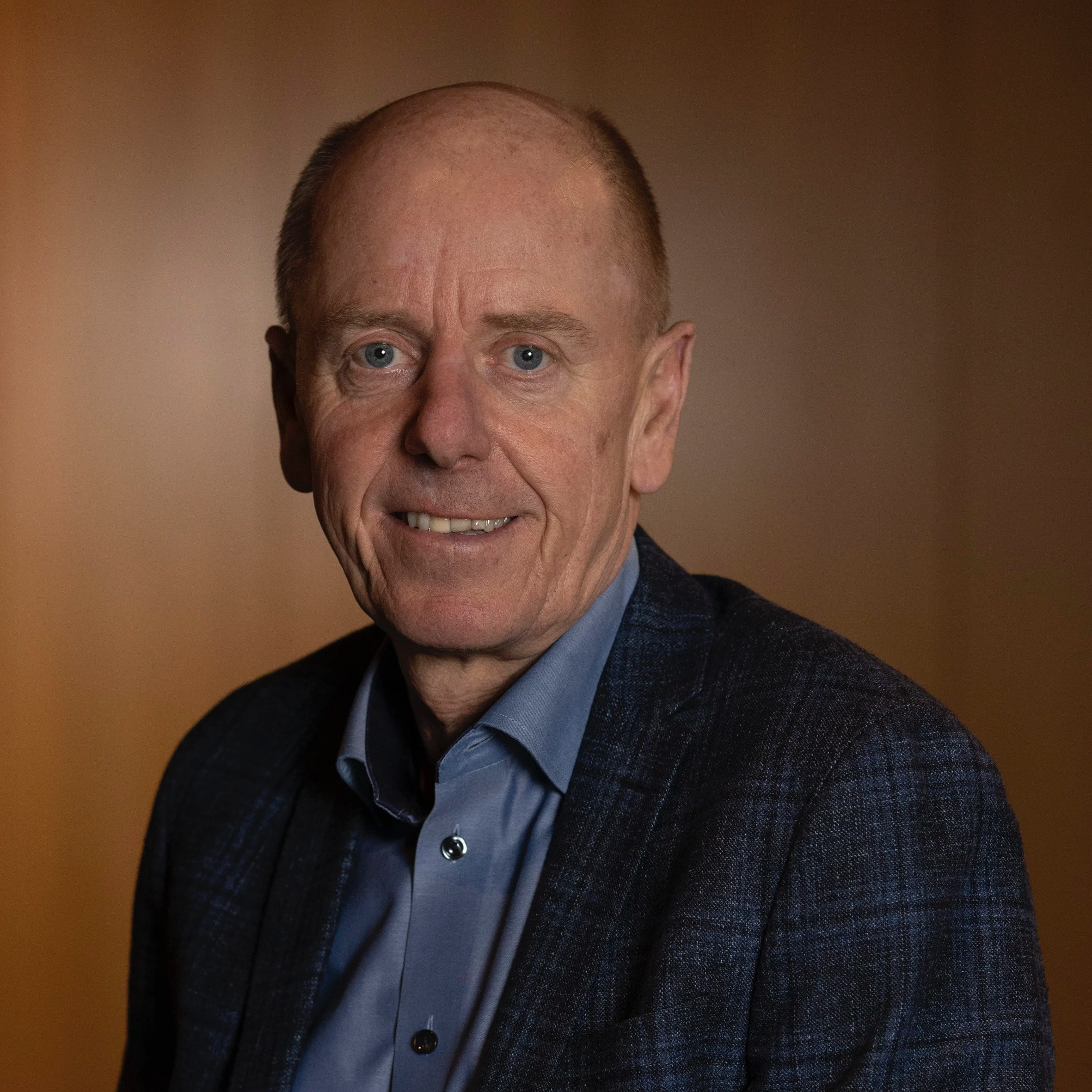 Headshot of an older man with short hair, blue eyes, smiling, wearing a blue button-up shirt and a dark blazer.
