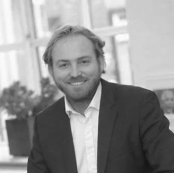 A man with blond hair and a beard, wearing a dark suit jacket and white shirt, smiling indoors with large windows and potted plants in the background.