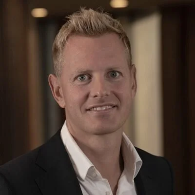 A man with light-colored hair and blue eyes, wearing a black suit jacket and a white shirt, smiling in an indoor setting.