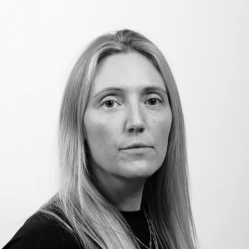A woman with long blonde hair wearing a black top, looking directly at the camera with a neutral expression, against a plain white background.
