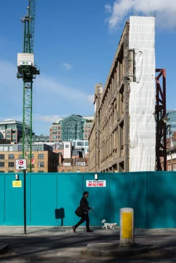 A person walking a small dog on a city sidewalk with a tall construction crane and partially covered building under construction in the background. The construction site is enclosed with a blue fence.