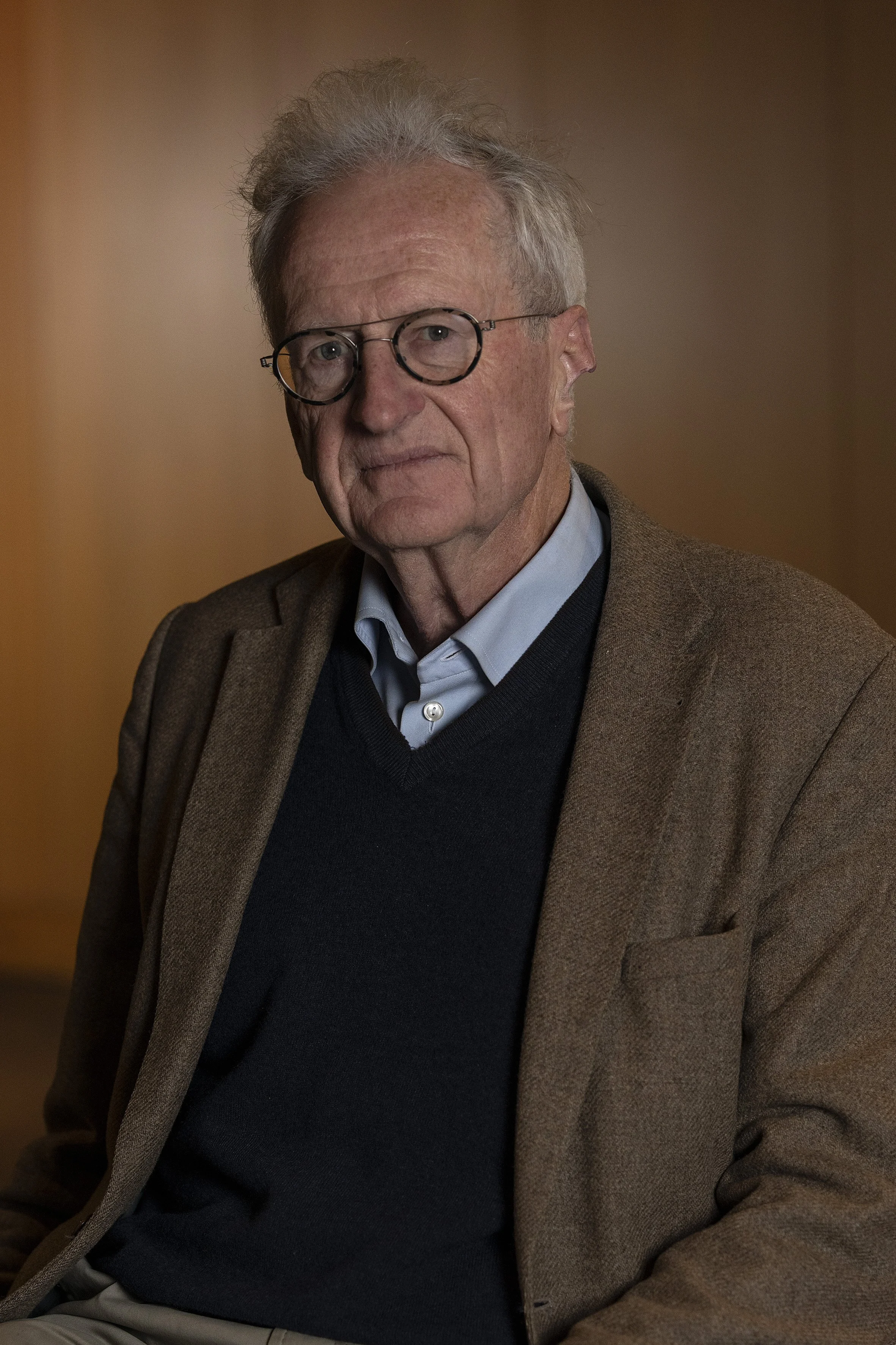 An elderly man with gray hair and glasses, wearing a blue shirt, black sweater, and brown blazer, sitting and looking at the camera against a warm, blurred background.