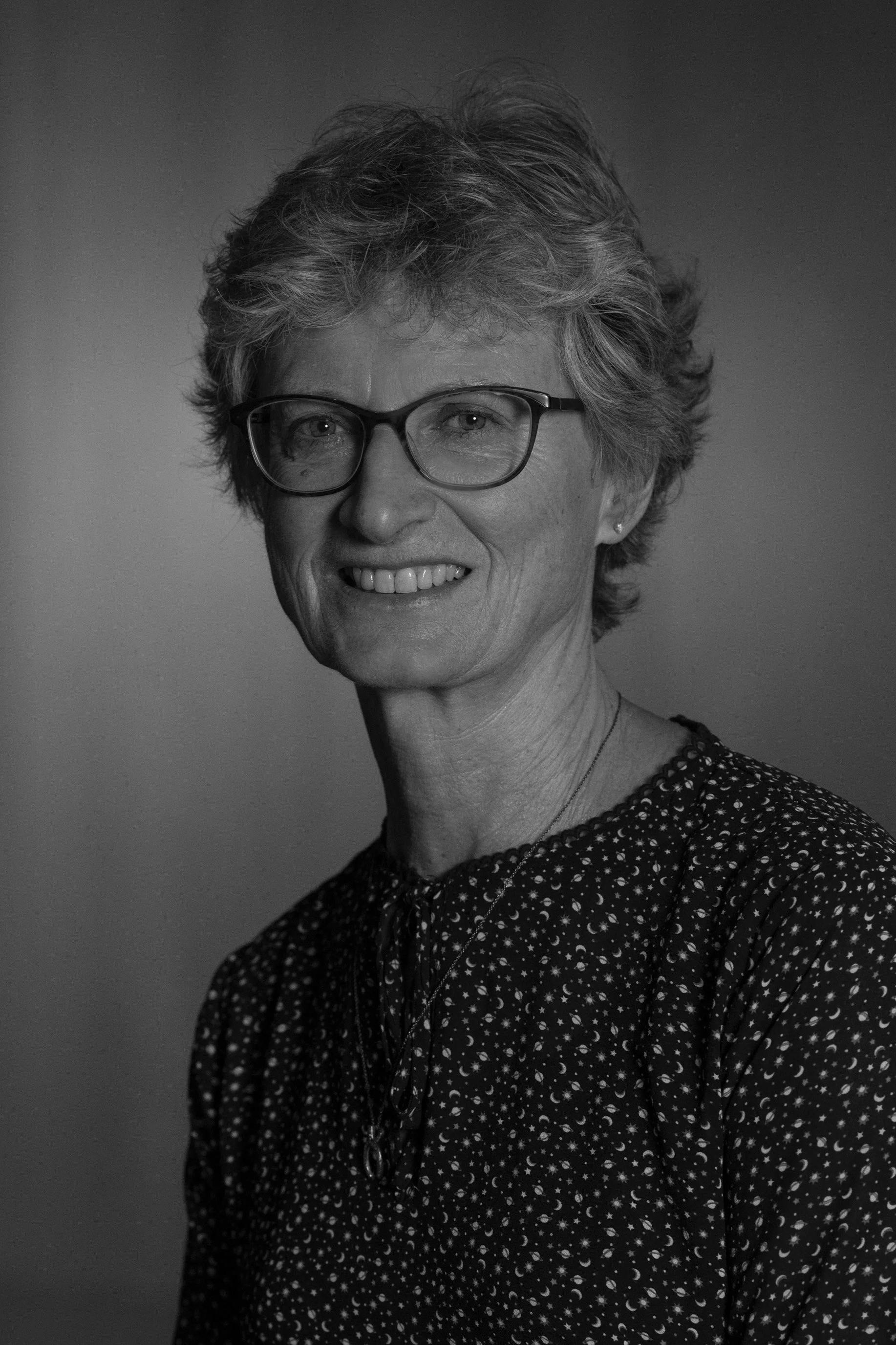 A woman with short gray hair, wearing glasses and a black blouse with small floral patterns, smiling at the camera against a warm brown background.