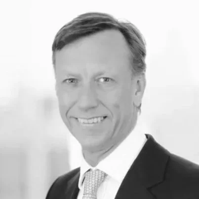 Portrait of a man in a dark suit with a light purple tie, smiling against a blurred office background.