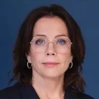 A woman with shoulder-length dark hair, glasses, and hoop earrings, wearing a dark top, against a dark blue background.