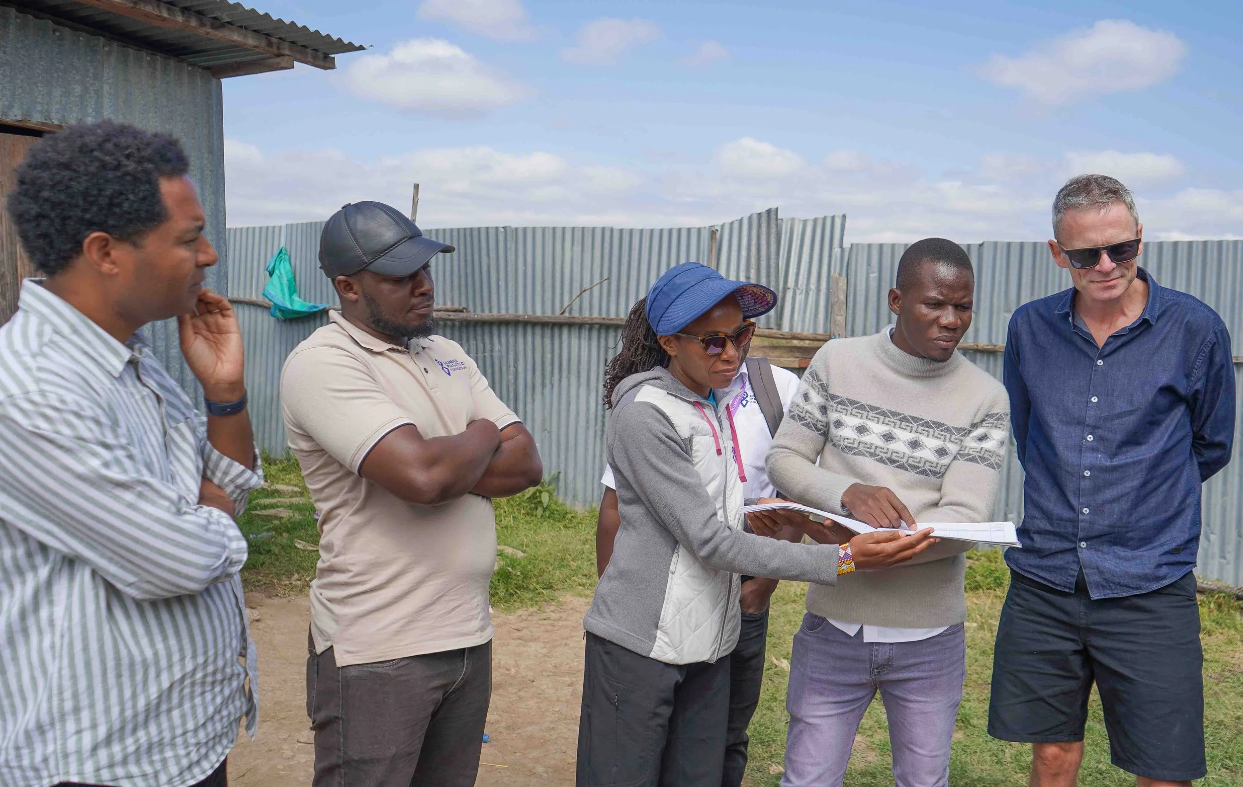Group of six people outdoors, some looking at a woman holding a notebook, with fencing and a blue sky in the background.