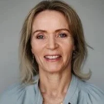 A woman with shoulder-length blonde hair smiling at the camera, wearing a light blue top against a plain background.