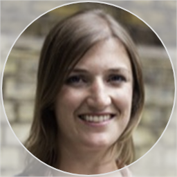 A smiling woman with long brown hair standing outdoors in front of a stone wall.