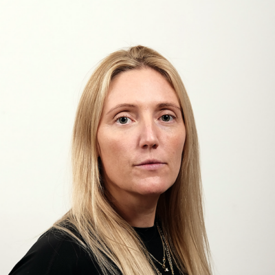 A woman with long blonde hair wearing a black top, looking directly at the camera with a neutral expression, against a plain white background.