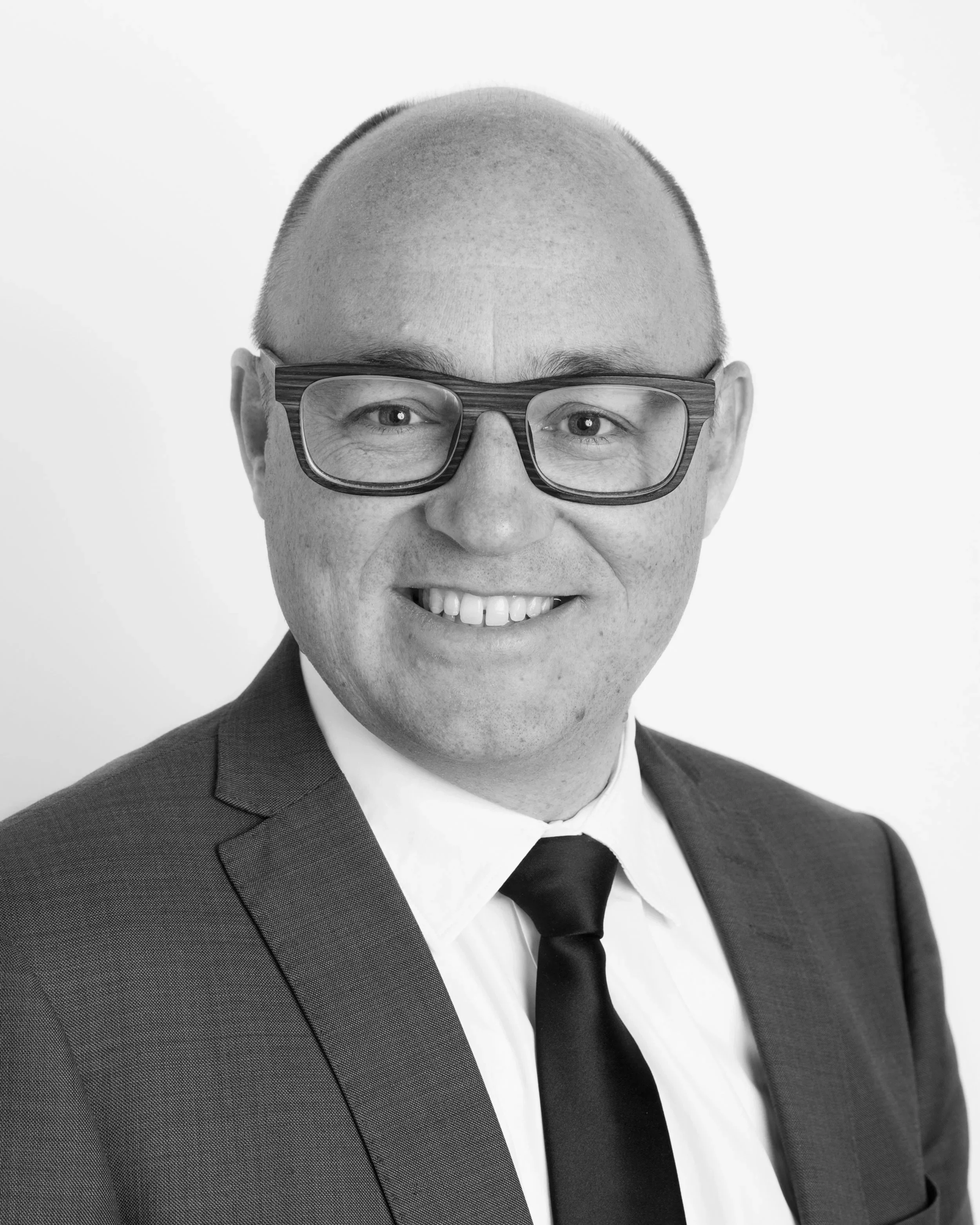 A man with glasses wearing a black suit, white shirt, and black tie, smiling against a plain white background.