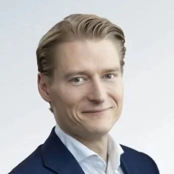 A professional headshot of a smiling man in a suit and tie against a textured blue background.