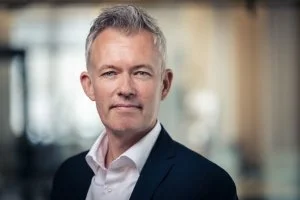A middle-aged man with short gray hair in a business suit, standing outdoors with a blurred city background.